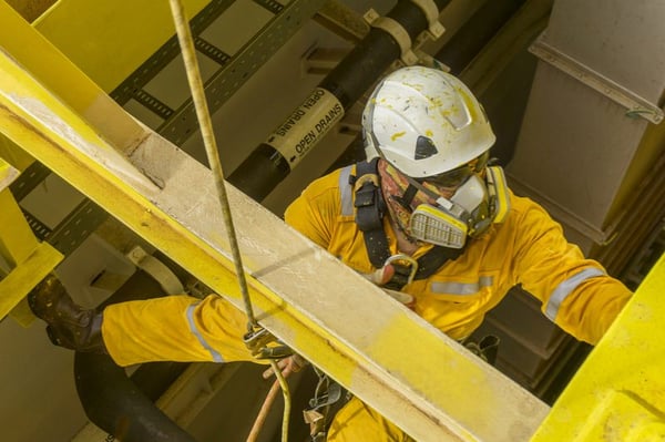 worker suspended next to a metal beam, wearing yellow coveralls, a white hard hat, and a full-face respirator mask