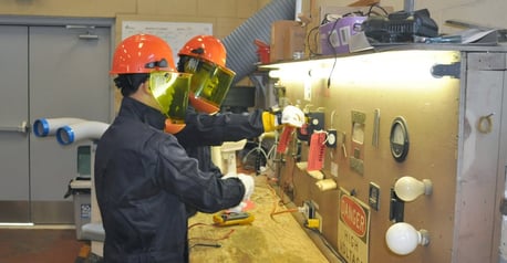 workers wearing hard hats, faceshields, coveralls, and protective gloves doing lockout-tagout training in front of a panel of gauges and switches