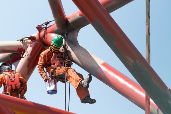 Worker wearing coveralls, hard hat, boots, and gloves suspended next to metal bridge structure