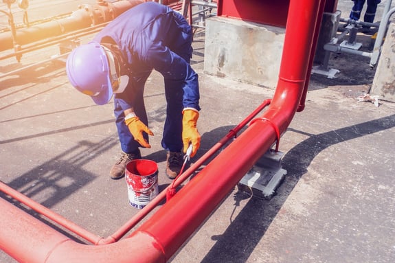 worker wearing coveralls, hard hat, gloves, and disposable respirator mask painting a large pipe red
