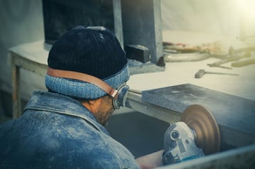Worker crouched at a work bench using a grinding tool to smooth a board. Dust covers the worker and the bench. Protect workers from silica exposure.