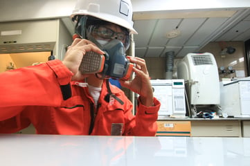 worker wearing orange coveralls, white hard hat, safety glasses, and donning a respirator mask